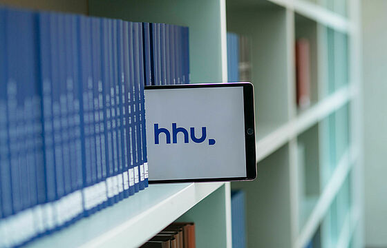 Ein Bücherregal mit blauen Bänden, zwischen denen ein Tablet steckt, auf dem das HHU-Logo zu sehen ist